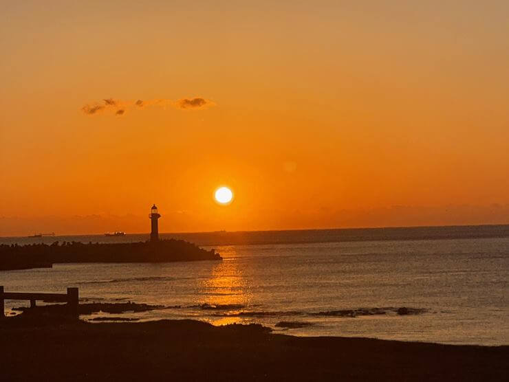 일출 사진으로 바다와 등대가 보이고 등대 뒤에서 해가 떠오르고 있다. 그리고 떠오르는 해 주변 하늘은 빨갛고 그 외는 아직 어둡다.
