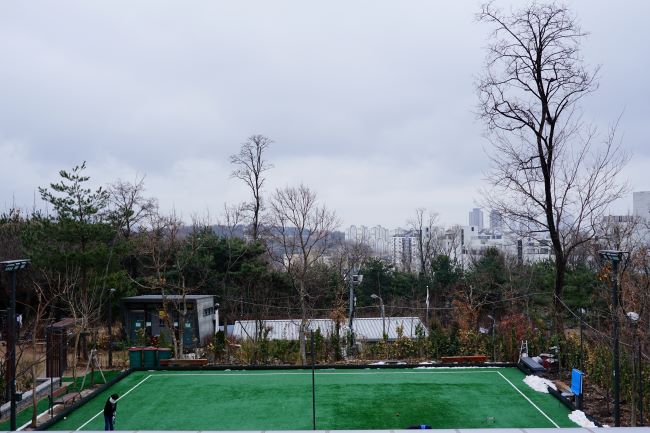 정자 아래에 보이는 파크골프장&#44; 녹색 마당&#44; 위로 보이는 잡목&#44; 흐린 하늘&#44; 검은 옷의 청소년(여자) 1&#44;