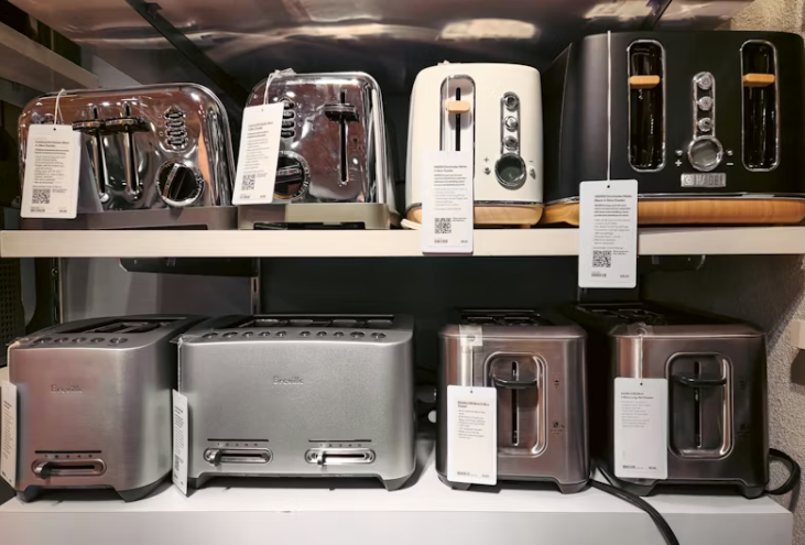 선반 위에 다양한 색상과 디자인의 토스터기들이 진열되어 있는 모습. 제품마다 라벨이 부착되어 있어 판매용 혹은 전시용임을 나타냄.