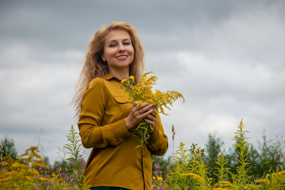 A cheerful woman stands in a field holding vibrant yellow wildflowers under a cloudy sky.