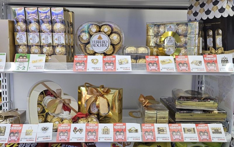 편의점 매대에 진열된 다양한 페레로 로쉐 초콜릿 선물 세트들과 그 앞에 꼼꼼하게 붙어있는 행사 가격표들. 여성 고객들이 인지도 있는 브랜드의 낱개 가격, 묶음 할인율, 그리고 통신사 할인까지 실시간으로 비교 분석하는 합리적이고 치밀한 구매 패턴의 배경.