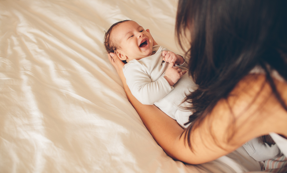 엄마가 침대에 누워 웃고 있는 아기를 안아주고 있다.