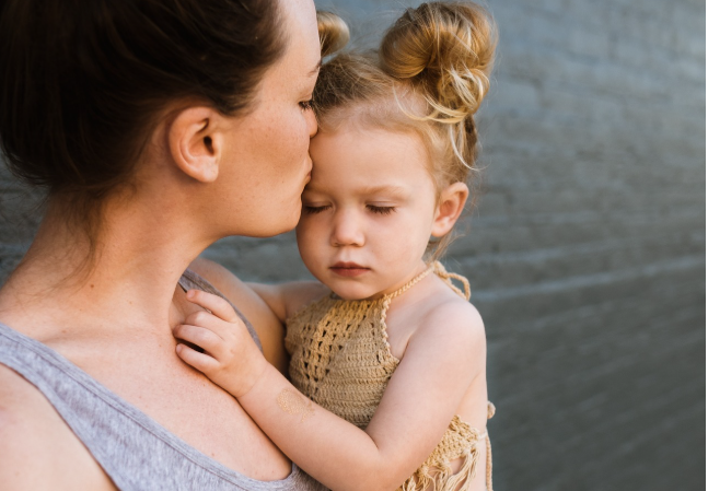 눈을 감고 엄마 품에 안긴 아이, 엄마는 아이의 이마에 부드럽게 입맞춤하며 감정을 다독이고 있다.