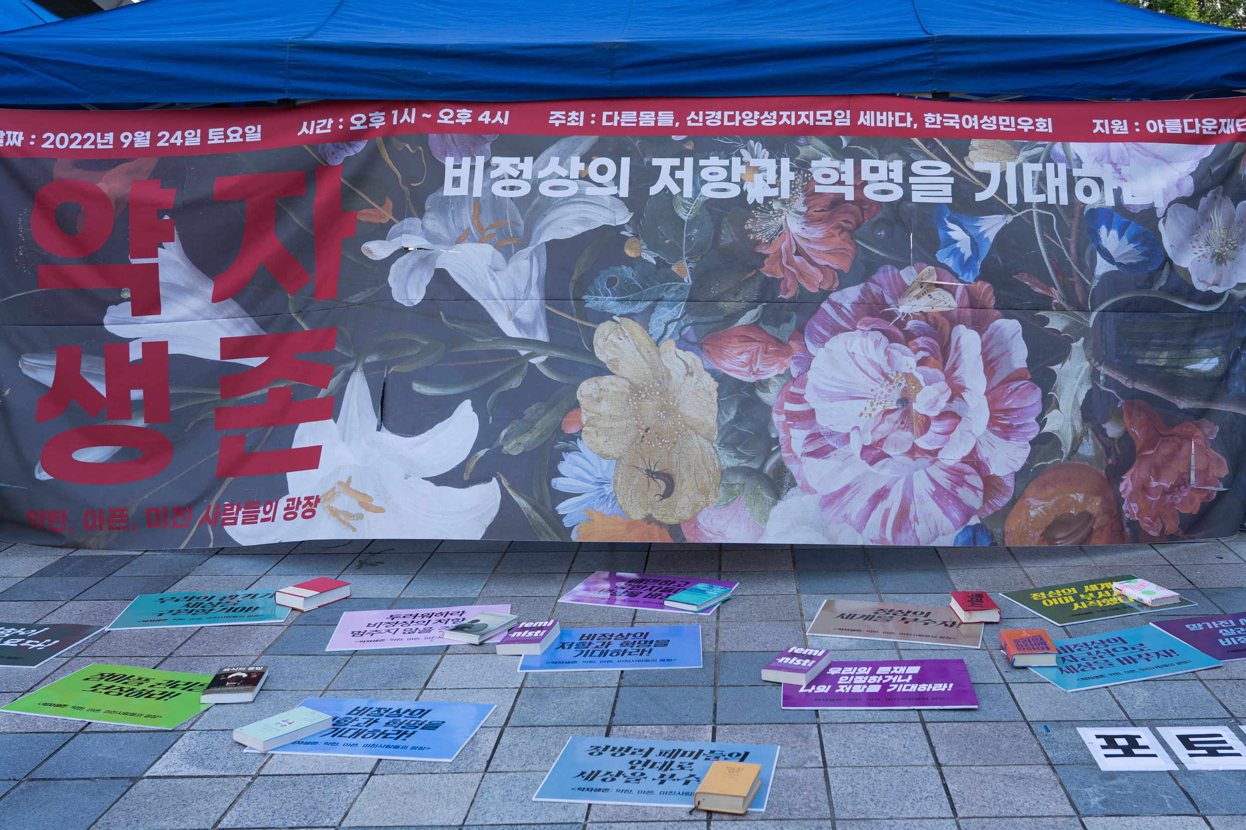 포토존에 <약자생존> 초대형 현수막이 걸려 있고, 바닥에는 구호가 적힌 피켓과 책이 있다.