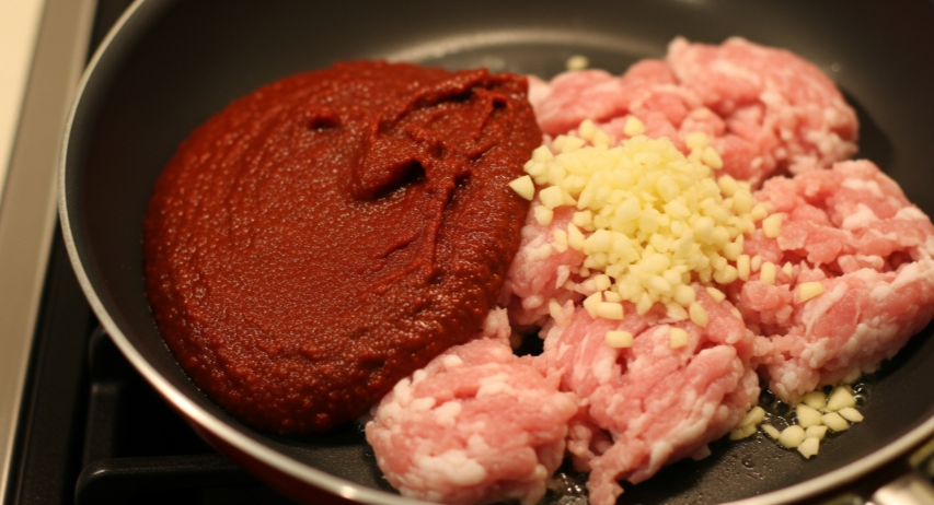 냄비에 다진 마늘과 함께 돼지고기를 볶고 있는 모습과 옆에 고추장, 고춧가루 등으로 미리 만들어 놓은 빨간색 양념장