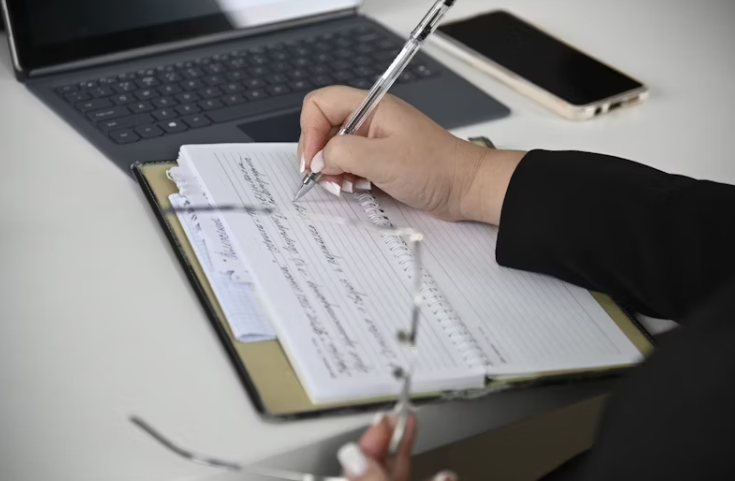 노트북 앞에서 공책에 메모를 남기는 직장인의 손, 한 손엔 펜을, 다른 손엔 안경을 들고 있는 모습