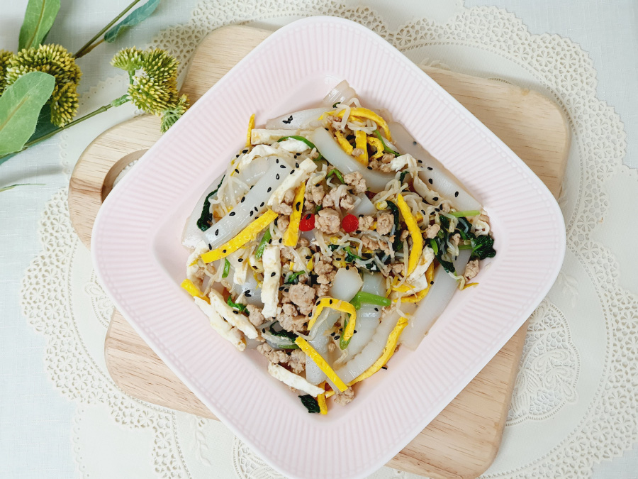 하얀 바닥 위에 네모난 나무 도마를 놓고&#44; 그 위에 네모난 유리 접시를 놓고 그 속에 돼지고기와 청포묵&#44; 계란지단&#44; 검은 깨&#44; 미나리&#44; 숙주나물을 넣고 버무린 탕평채를 담아 두고 찍은 사진