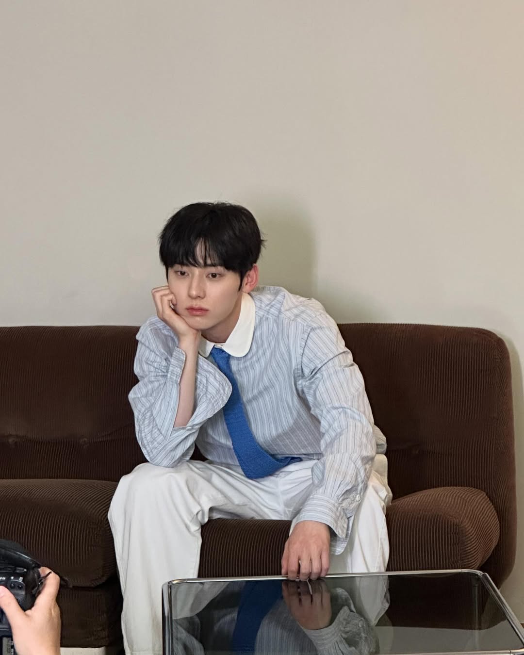 스프라이트 셔츠에 블루 넥타이를 매고 흰 바지를 입은 코디에 앉아서 턱을 괴고 있는 워너원 황민현의 사진