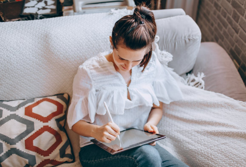 여성이 소파에 앉아 태블릿으로 무언가를 작성하고 있는 모습, 비대면 복지 신청을 연상케 하는 장면