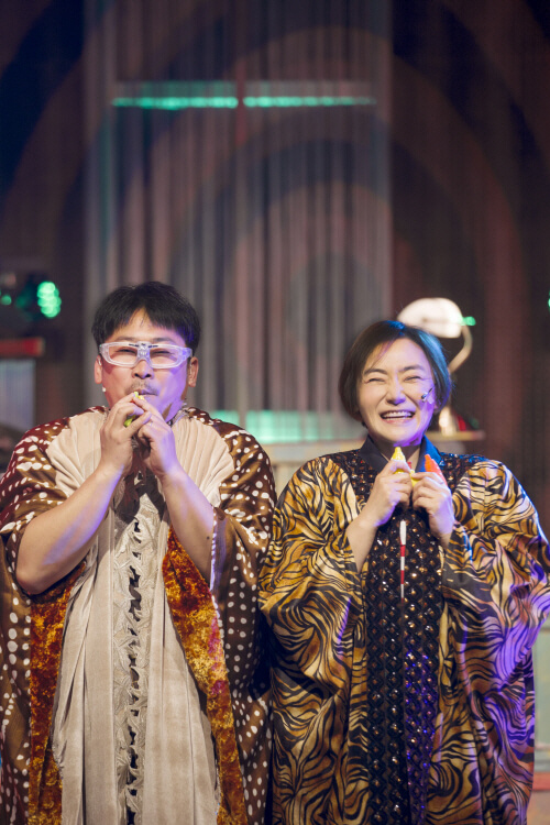 공연 중 개성 넘치는 의상을 입은 남녀 배우가 나란히 서서 플루트를 불고 있는 장면, 밝은 조명 아래 웃으며 관객과 교감함