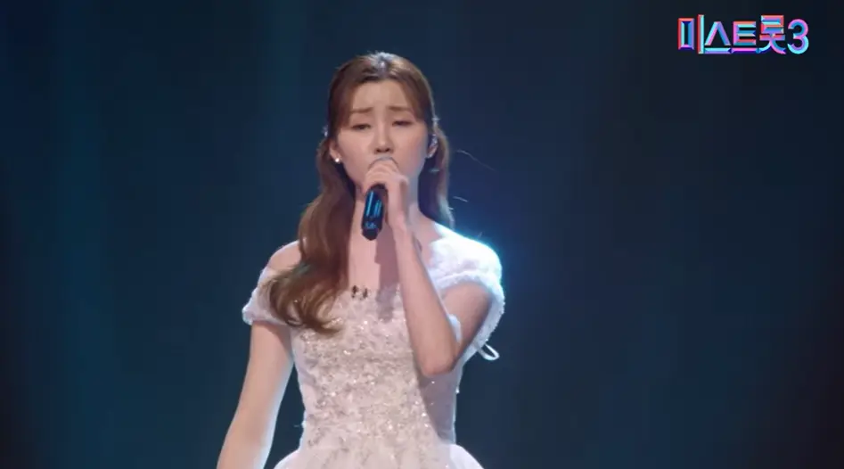 미스트롯3- 검푸른 배경 흰드레스를 입고 왼손으로 마이크를 든채 긴머리를 늘어뜨리고 노래하는 나영