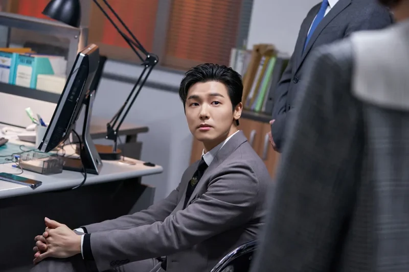 어느 사무실 책상앞에 회색 정장을 입고 다리 꼬면서 앉아 있는 강민혁