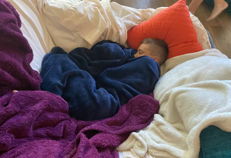 따뜻한 담요에 둘러싸여 바닥에서 편안하게 잠든 어린아이의 모습. 다양한 색상의 포근한 이불이 난방 없이도 따뜻한 실내 분위기를 보여줌.