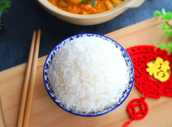 나무 도마 위에 하얀 쌀밥이 담긴 도자기 밥그릇이 있다. 왼쪽에는 나무 젓가락이 있고 뒤에는 찌개가 담긴 냄비가 있다.