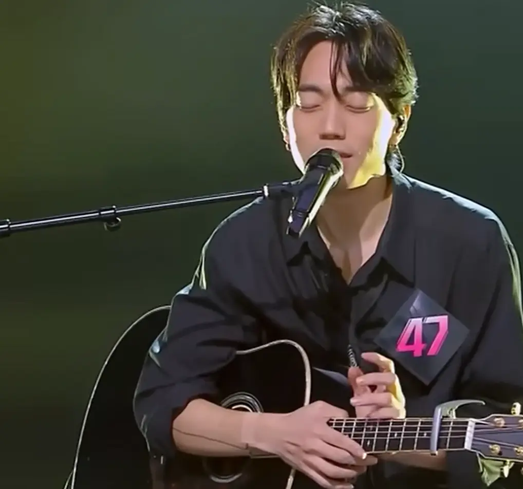 싱어게인3- 녹색배경 검은 셔츠 가슴에 47번호표를 달고 검은색 기타손으로 감싸안으며 스탠딩마이크를 앞에 두고 두눈 감은채 노래하는 47호 