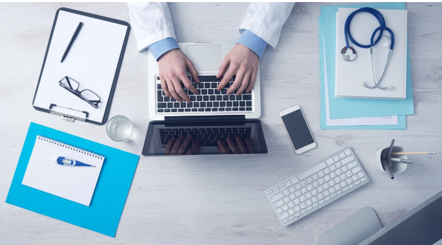 노트북 앞에서 보험 관련 서류를 검토 중인 책상 위 모습. 서류, 청진기, 안경, 계산기 등이 함께 놓여 있다.