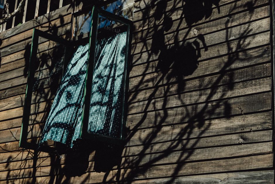 Open window on a wooden house with patterned curtains and tree shadows, creating a rustic look.