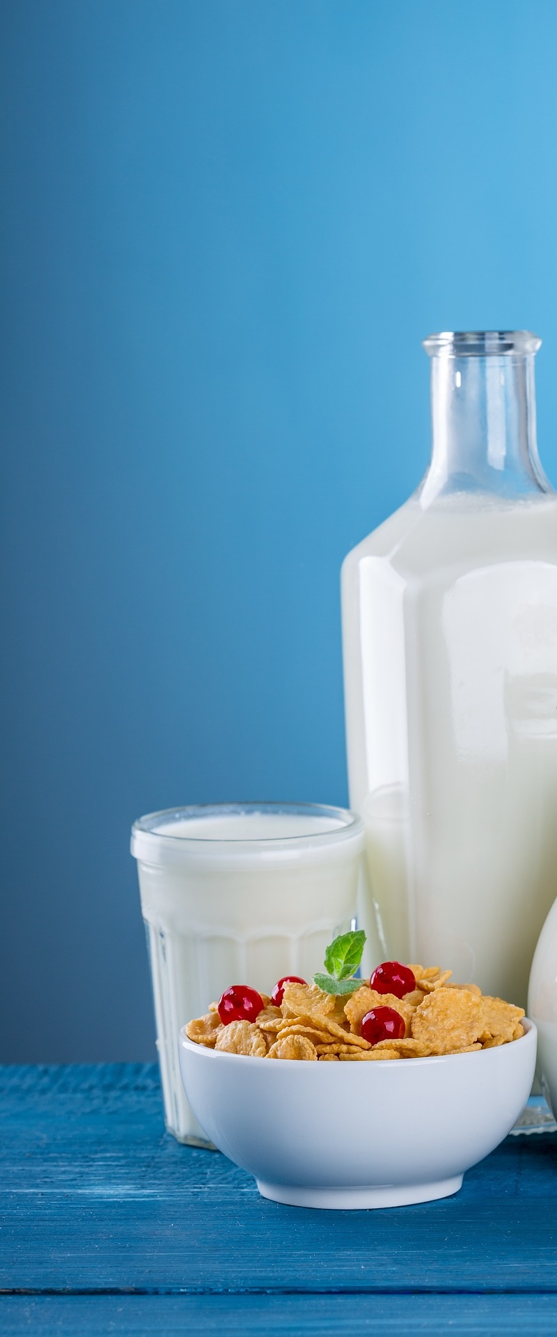 파란색 배경 앞에 우유가 가득 담긴 유리병과 유리 저그, 그리고 빨간 베리가 올라간 시리얼 보울이 놓여 있는 모습