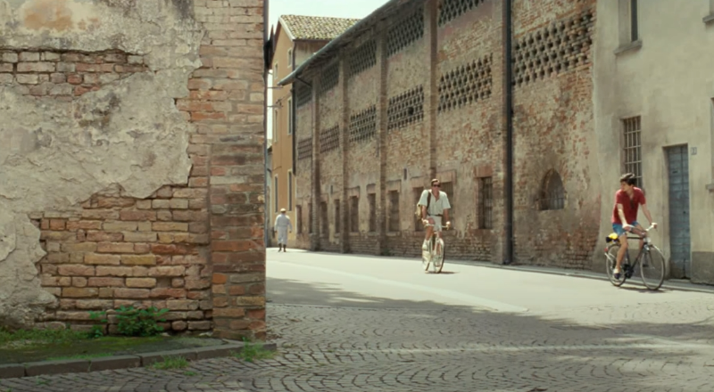 벽돌이 있는 건축물이 있고 남자 둘 이 자전거를 타고 있다.