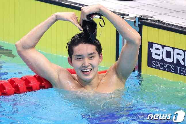 김우민 선수가 수영장 풀 안에서 팔로 크게 하트를 그리며 웃고 있다
