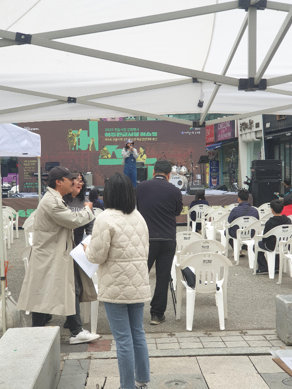 공연장에서는 사회자가 사회를 보고 있고 뒷편에서는 공연 관계자들이 이야기 중이다. 의자에 관중은 별로 없다.