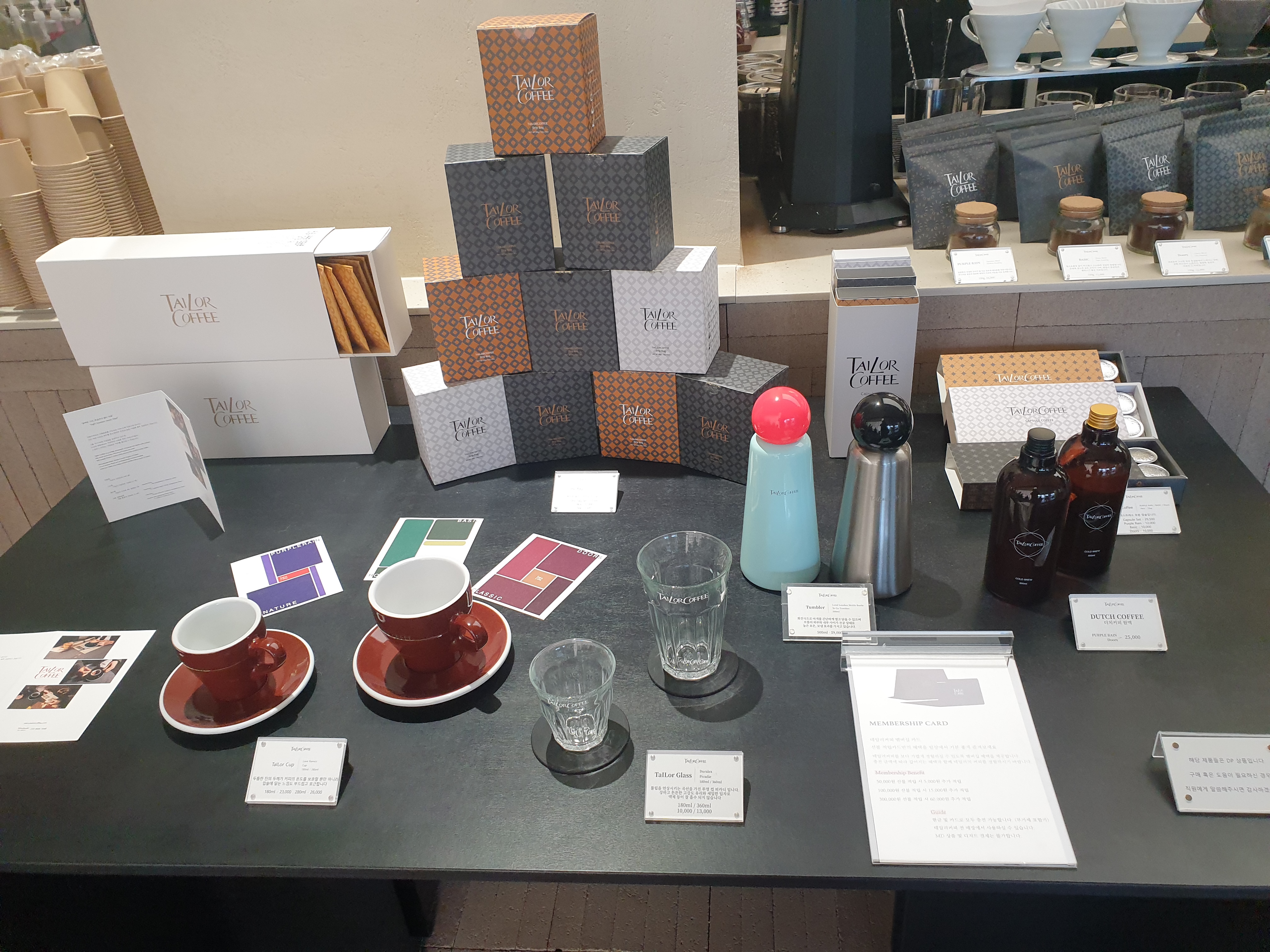 굿즈 진열대. 검은색 테이블 위에 컵&amp;#44; 잔&amp;#44; 텀블러와 함께 캡슐&amp;#44; 드립백이 보인다.