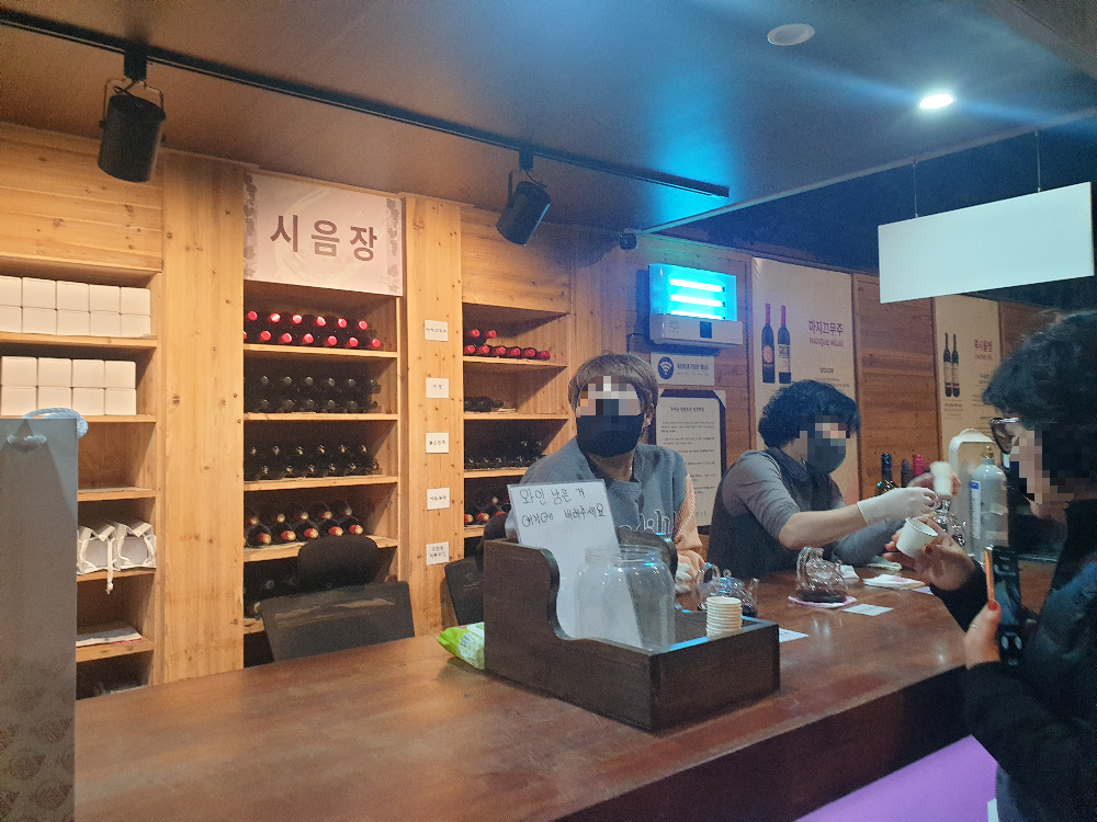 시음장에 3명이 시음을 도와주고 있음. 손님들이 줄을 서서 시음 중