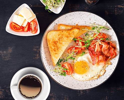 둥그런 접시에 삼각형의 토스트 2개와 달걀프라이, 베이컨, 야채가 있다. 주변엔 하얀 치즈와 잘려진 방울토마토가 담긴 그릇이 있다.