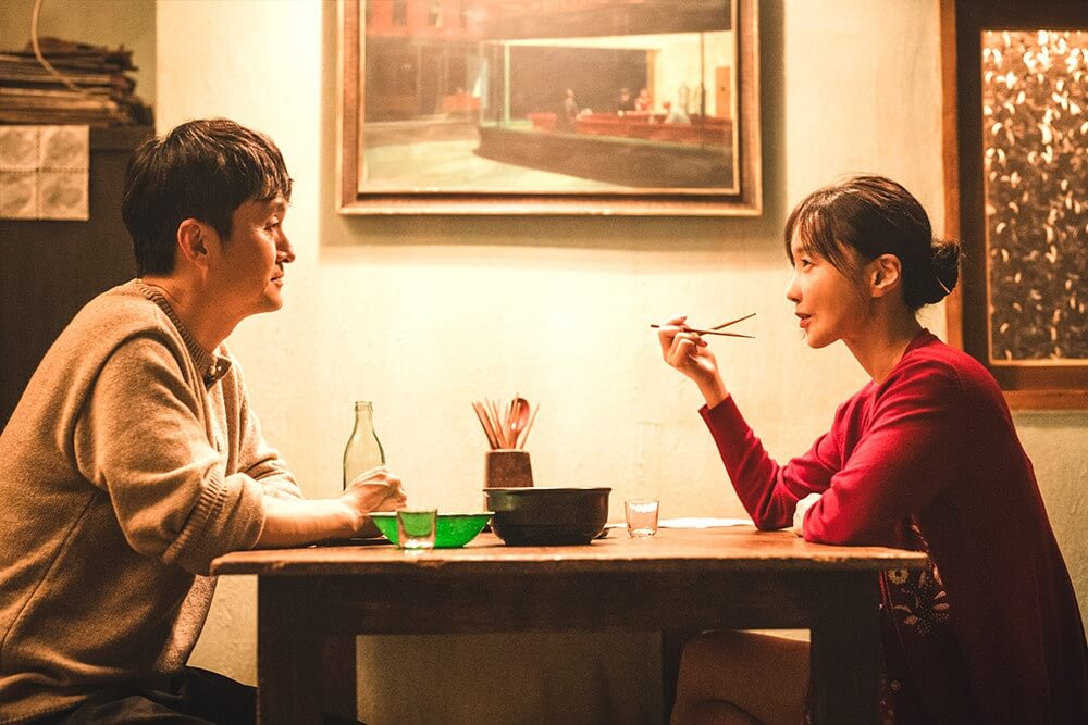 영화 더 킬러스 스틸컷 &ndash; 두 인물이 식탁을 사이에 두고 대화를 나누며 미묘한 긴장감을 형성하는 장면