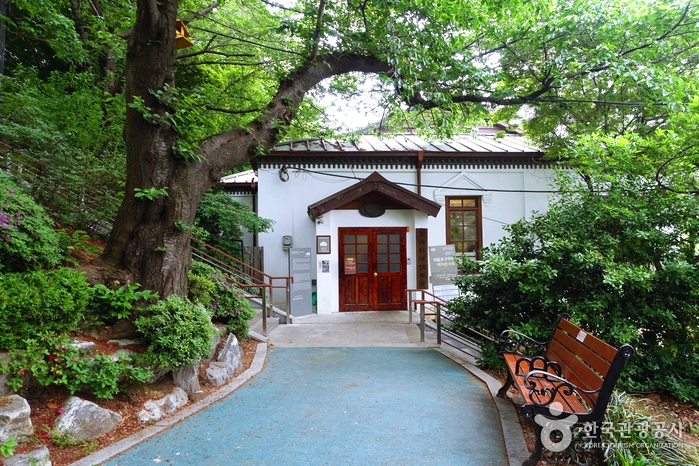 하얀건물에 지붕을 양철로 덮은 집한채가 있고 양쪽에 푸른 나무들이 있다.