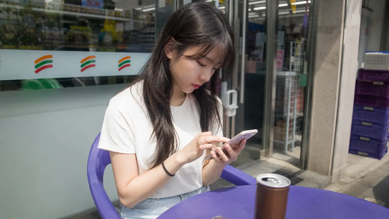 편의점 야외 테이블에 앉아 스마트폰으로 핀테크 앱의 합법적인 포인트 전환 과정을 집중해서 따라 하는 20대 여성