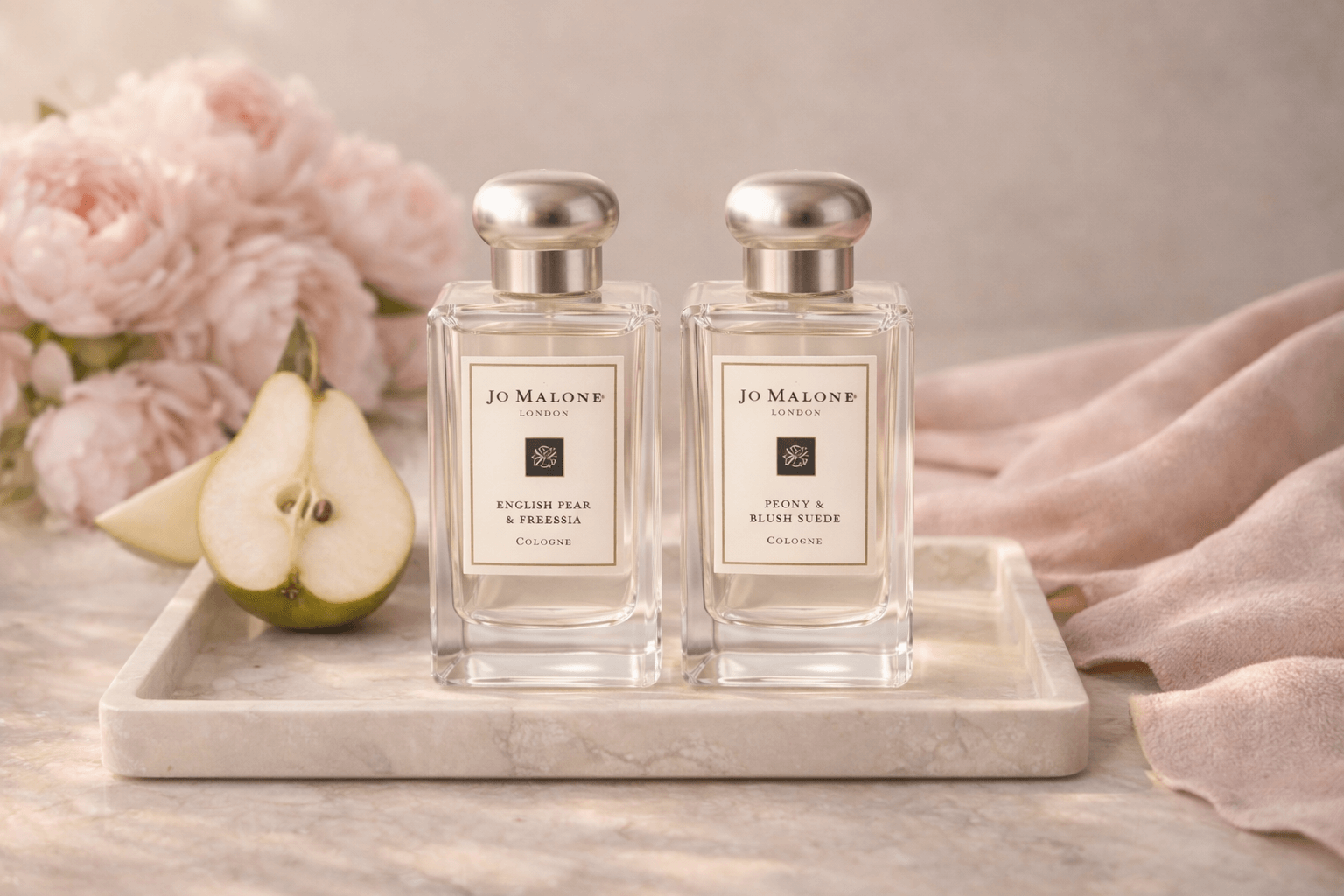 대리석 트레이 위에 나란히 놓인 조말론 피오니 앤 블러쉬 스웨이드와 잉글리쉬 페어 앤 프리지아 향수병, 배 슬라이스와 작약 꽃, 스웨이드 패브릭으로 연출한 우아한 레이어링 퍼퓸 이미지