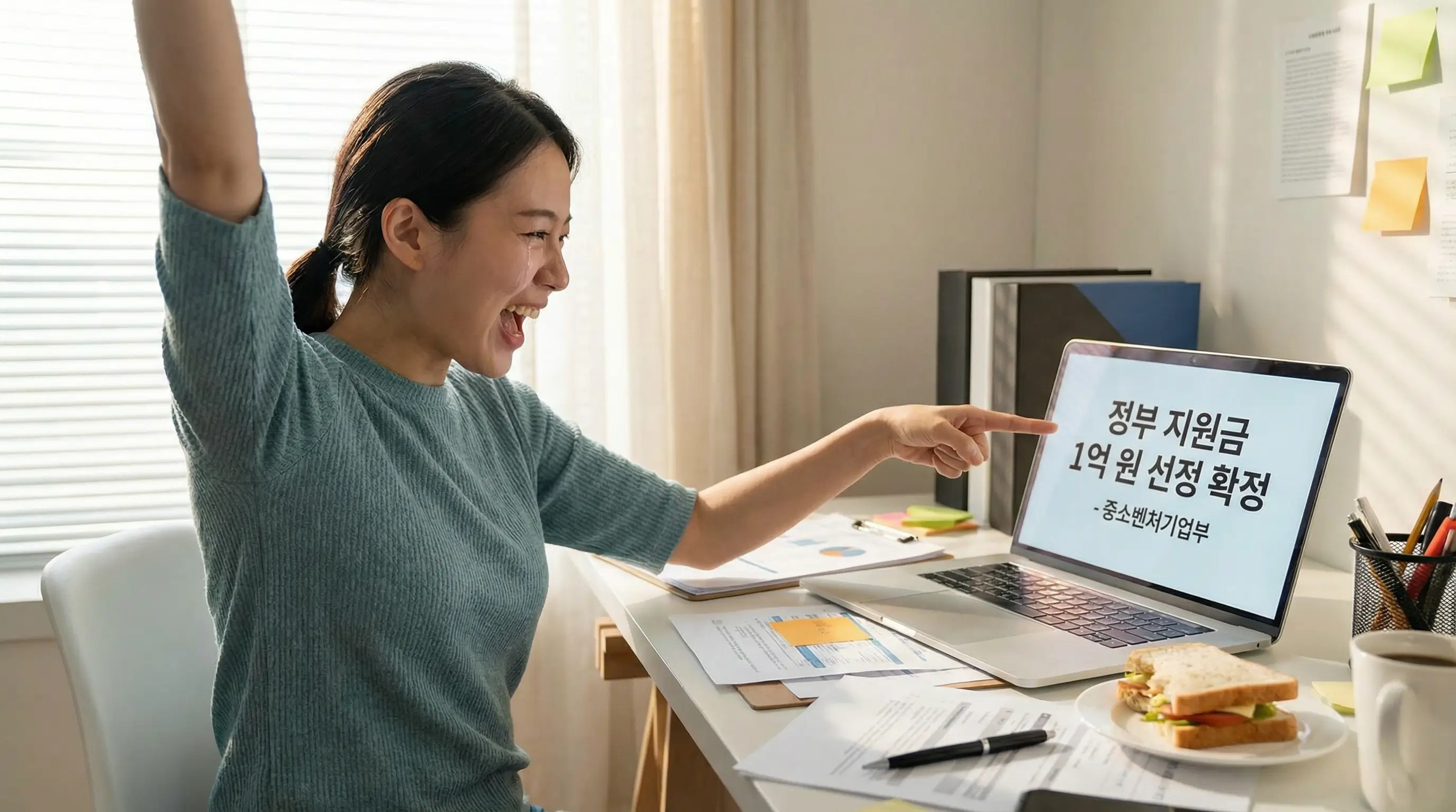 노트북 앞에서 사업계획서를 작성하며 정부 지원금 1억 원 선정 소식을 듣고 기뻐하는 예비 창업자의 모습