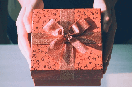 사각형의 연한 다홍색 선물상자가 있고 두 손이 선물상자를 들고 전해주는 장면이다.