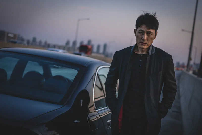 자동차를 배경으로 자켓을 입고 걷고 있는 영화 보호자의 수혁