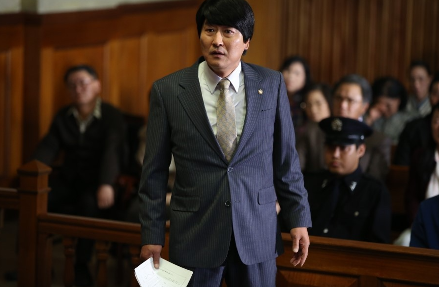 법정에서 서류를 손에 들고 서 있는 송우석의 모습, 뒤로 방청객과 경찰관이 보이는 1980년대 법정 장면