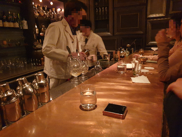 따뜻한 우드 톤의 카운터에 셰이커들이 정렬되어 있다. 바텐더 2명이 칵테일을 제조 중이다.