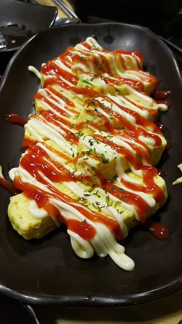 검은색 접시위에 두꺼운 계란말이 위로 마요네즈 케찹이 뿌려진 계란말이