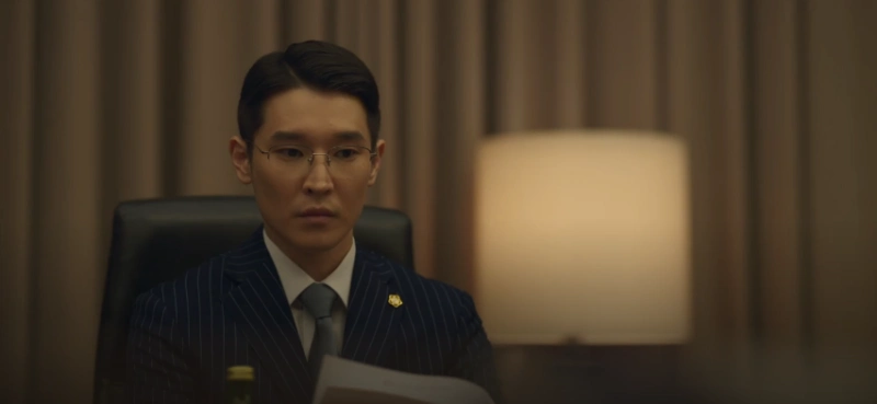 커튼이 있는 회의실에 단정히 빗어 넘긴 머리와 파랑색 고급 슈트를 입고 앉아 있는 유괴의 날 변호사 최택균 캐릭터