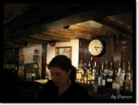 영국 런던 근교의 한 펍 내부. 나무 들보가 드러난 낮은 천장 아래, 술병들이 진열된 바 뒤편 벽에는 둥근 시계가 걸려 있고, 바텐더로 보이는 여성이 어슴푸레한 조명 아래 서 있다. 따뜻하고 아늑한 분위기가 느껴진다.