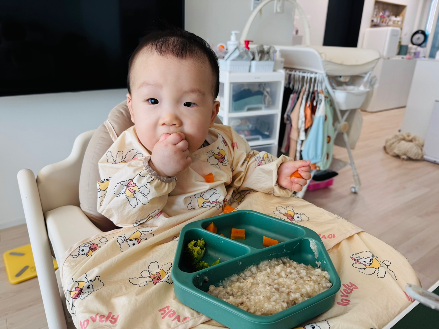 당근을 한입 먹으며 브로콜리와 죽이 담긴 식판 앞에 앉아 있는 찬솔이