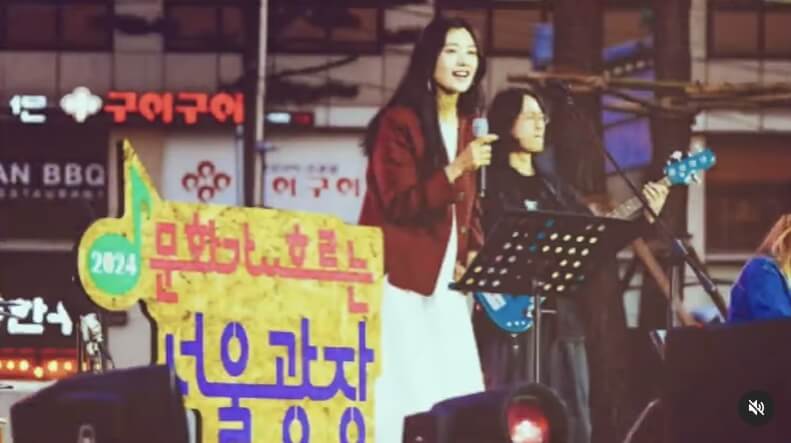 문화가 흐르는 서울광장 무대에서 마이크를 든 여성 가수가 밴드와 함께 라이브 공연을 펼치는 장면, 무대 앞 2024 행사명 간판 포함
