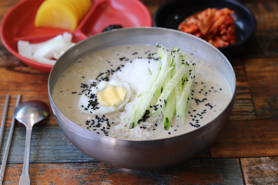나무 식탁 위에 오이오 계란이 고명을 올려진 시원한 콩국수를 담은 양은그릇과 김치를 담은 검은 그릇, 그리고 단무지와 양파, 춘장을 담은 빨간 그릇을 놓고 찍은 사진