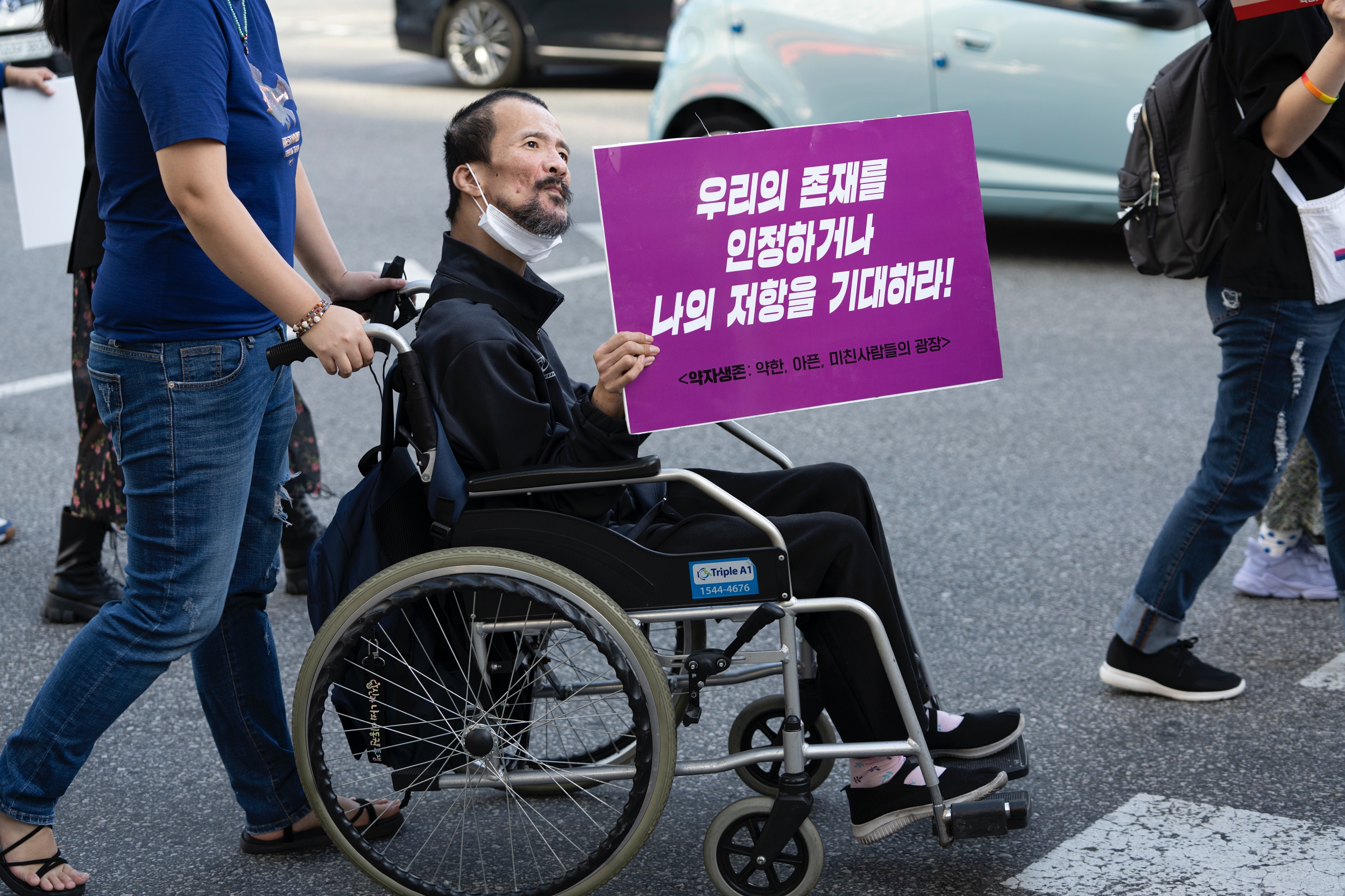 휠체어를 탄 시민이 '우리의 존재를 인정하거나 나의 저항을 기대하라!'라는 피켓을 들고 행진하고 있다.