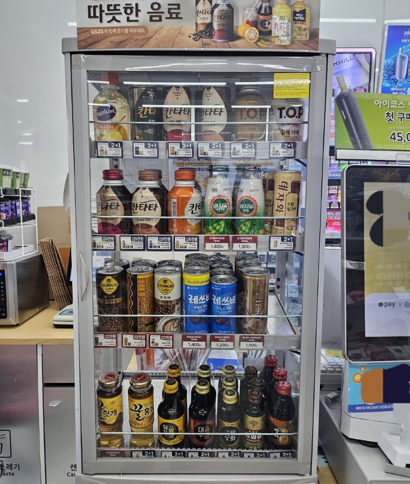 편의점 카운터 옆에 설치된 온장고 내부 모습. 캔커피와 병음료들이 정갈하게 진열되어 있으며, 사장이 데이터에 기반해 재고 수량을 조절하며 관리하는 운영의 디테일을 보여주는 사진.