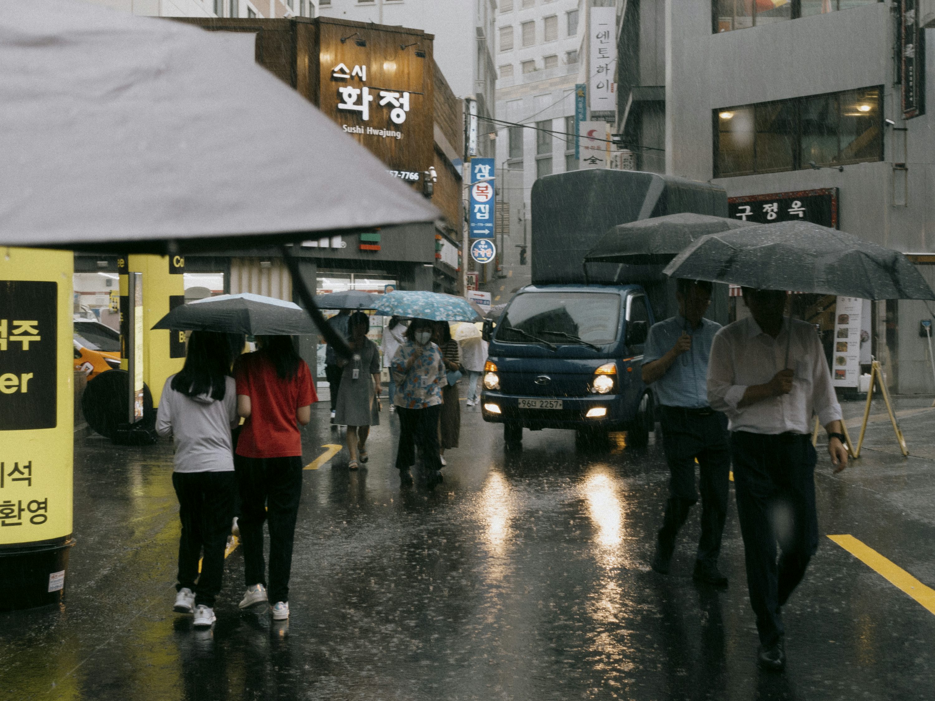 한국 도심 거리에서 폭우가 쏟아지는 가운데 여러 사람들이 우산을 쓰고 걸어가고 있으며, 가게 간판과 차량 불빛이 빗속에 반사되는 장마철 풍경
