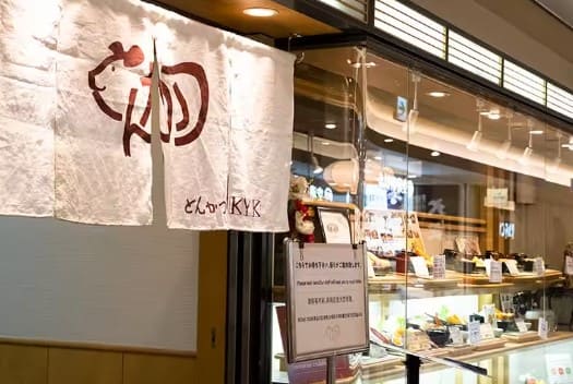 노렌(일본식 커텐)이 가게 위에 붙어 있고 투명한 진열대 안에 음식들이 진열되어 있는 가게 외부 사진