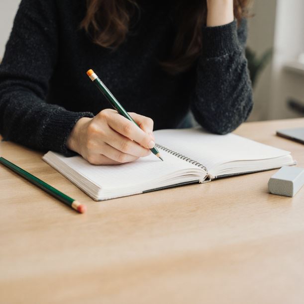 책상에 앉아 노트에 글을 쓰고 있는 사람의 모습. 연필과 지우개도 함께 놓여 있다.