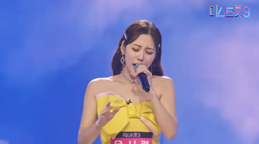 미스트롯3- 가슴에 커다란 노란리본이 달린 민소매 원피스에 배부분 분홍색 바탕 흰글씨의 윤서령이 적힌 이름표를 왼손으로 마이크를 잡고 눈감은채 애절하게 노래하는 윤서령