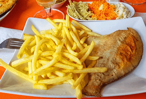 네모난 하얀 접시에 가자미 튀김 한 마리와 감자튀김이 가득 있다. 식탁 주변에는 맥주 잔과 야채 튀김이 있다.